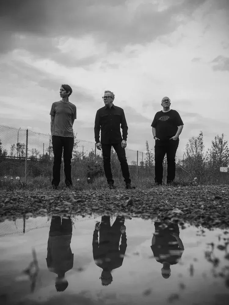 Three-piece band standing in a gravel lot, mirrored in a rain puddle