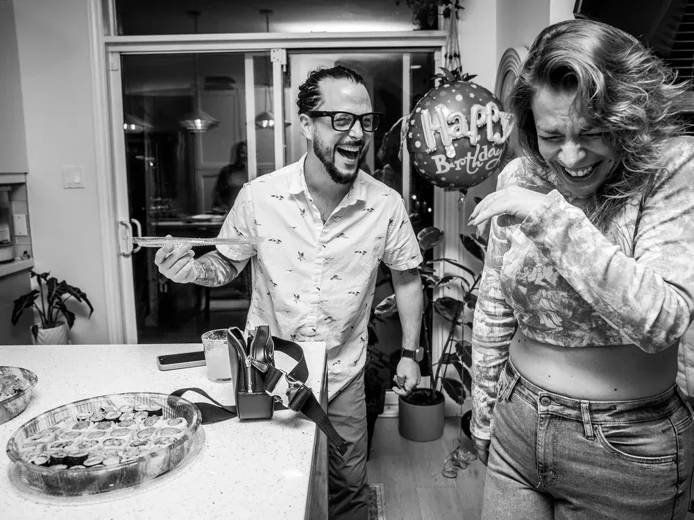 Couple mid-laugh in a kitchen at a birthday party, balloon behind them