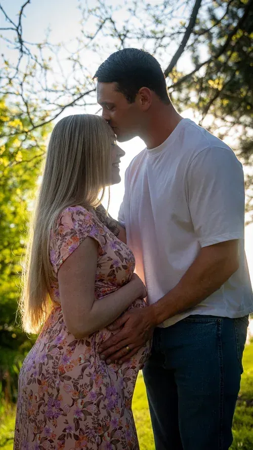Expecting father kissing his partner's forehead under backlit trees