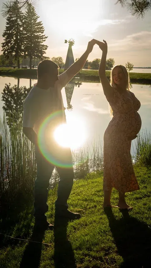 Expecting couple dancing by a lake, sun flaring between them