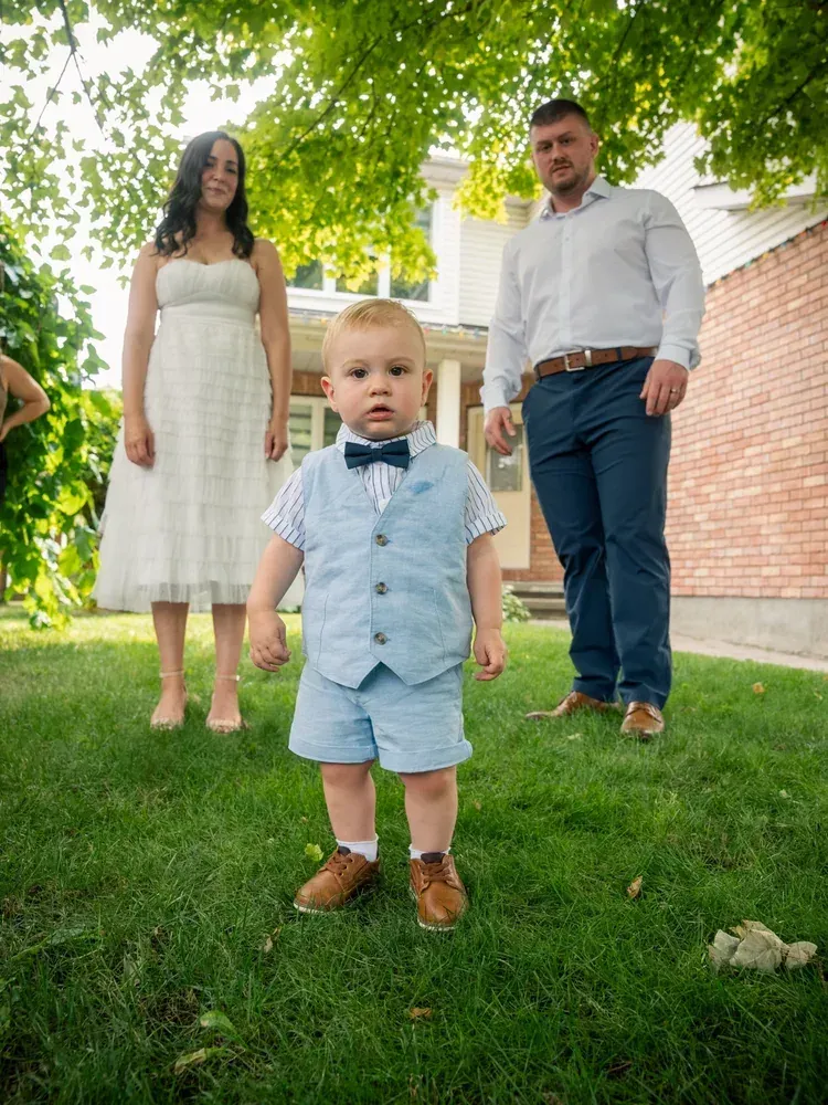 Toddler in a bow tie and blue suit walking ahead of his parents on the lawn