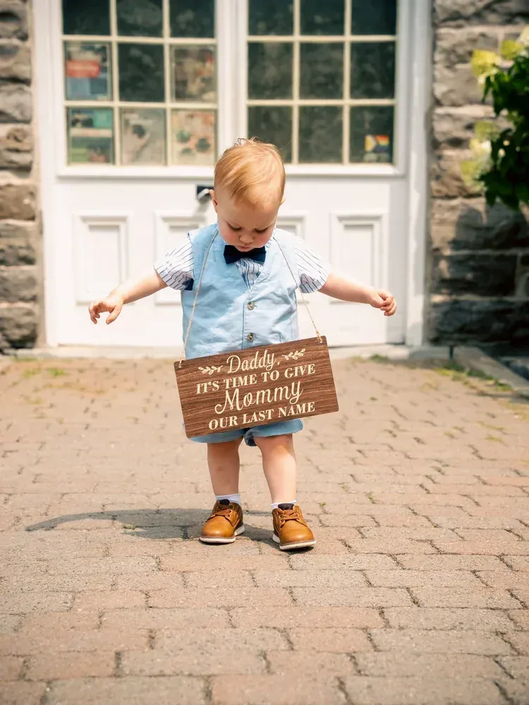 Toddler carrying a wooden sign reading 'Daddy, it's time to give Mommy our last name'