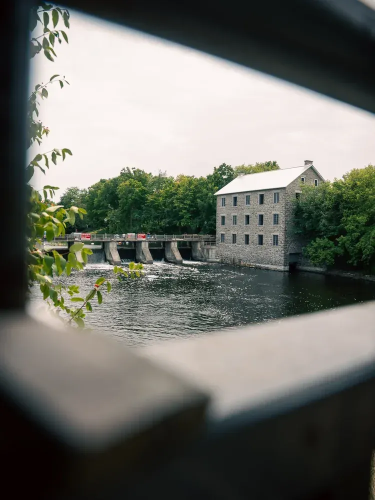 A stone grist mill and its dam framed through the gap in a wooden bridge railing